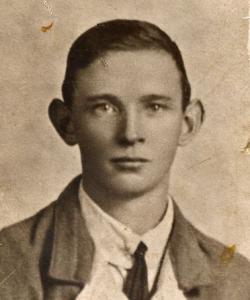 Posed sepia photograph, head and shoulders of James Nolan prior to enlisting aged 20. Freshfaced, short dark hair, wearing a white shirt with narrow knotted tie and a lighted coloured relaxed jacket.